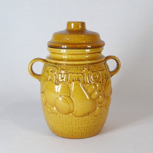 Puede incluir: Un tarro de cerámica amarillo con tapa, que presenta un patrón tejido y diseños de frutas en relieve. El tarro tiene dos asas y la palabra "Rumtopf" está escrita en la parte delantera.