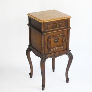 Puede incluir: Una mesita de noche de madera oscura con una tapa de mármol, un cajón y una puerta de armario con un diseño tallado. La mesita de noche tiene cuatro patas cabriole.