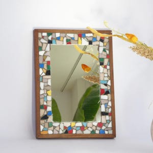 May include: A rectangular mirror with a mosaic tile border in various colours, including red, blue, yellow, and white. The frame is made of wood. The mirror reflects a room with a green plant and dried flowers.