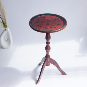 Puede incluir: Una mesa pedestal redonda de color rojo con borde negro y motivos florales pintados a mano en el tablero y la base de tres patas. La mesa presenta una columna central torneada y delgada con tres patas curvas, decoradas con motivos botánicos.
