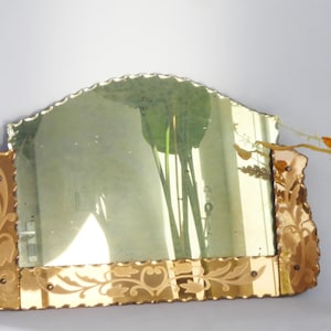 Op de afbeelding: Een antieke spiegel met een geschulpte bovenrand en een decoratieve gouden lijst. De lijst heeft geëtste bloemmotieven en een getextureerde, onregelmatige rand. De spiegel reflecteert een groene plant.