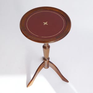 Op de afbeelding: Een kleine, ronde houten tafel met een rood leren blad en een gouden stervormig ontwerp. De tafel heeft drie poten en een geflute kolom.