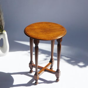 May include: A round wooden side table with a warm brown finish and a visible wood grain pattern on the top surface. The table features four slender, turned legs connected by a central cross-shaped stretcher base, providing structural support.