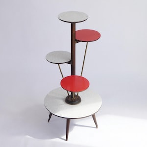 May include: A mid-century modern plant stand with a dark brown central pole and multiple circular shelves. The shelves alternate between red and white speckled surfaces, supported by thin metal rods and wooden legs.