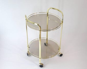 May include: A gold metal serving cart with two glass shelves. The cart has a round top and four wheels.
