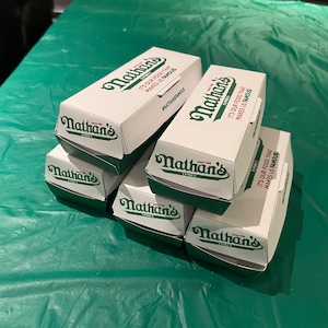 K&ouml;nnte beinhalten: Ein Stapel aus sechs wei&szlig;en Pappkartons mit gr&uuml;ner Beschriftung. Die Kartons sind mit "Nathan's Famous" beschriftet und es steht "It's our food that makes us famous" darauf.