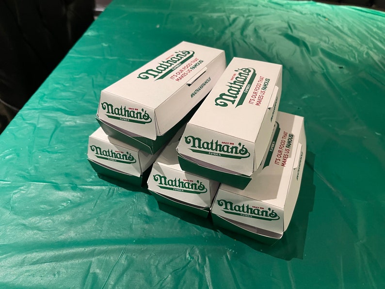 K&ouml;nnte beinhalten: Ein Stapel aus sechs wei&szlig;en Pappkartons mit gr&uuml;ner Beschriftung. Die Kartons sind mit "Nathan's Famous" beschriftet und es steht "It's our food that makes us famous" darauf.