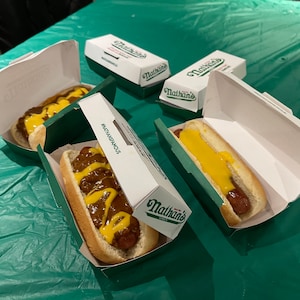 K&ouml;nnte beinhalten: Drei Nathan's Famous Hotdogs in wei&szlig;en Papierbeh&auml;ltern mit gr&uuml;nen Deckeln. Zwei Hotdogs sind mit Chili und gelbem Senf belegt. Ein Hotdog ist mit gelbem Senf belegt. Die Hotdogs befinden sich auf einer gr&uuml;nen Tischdecke.