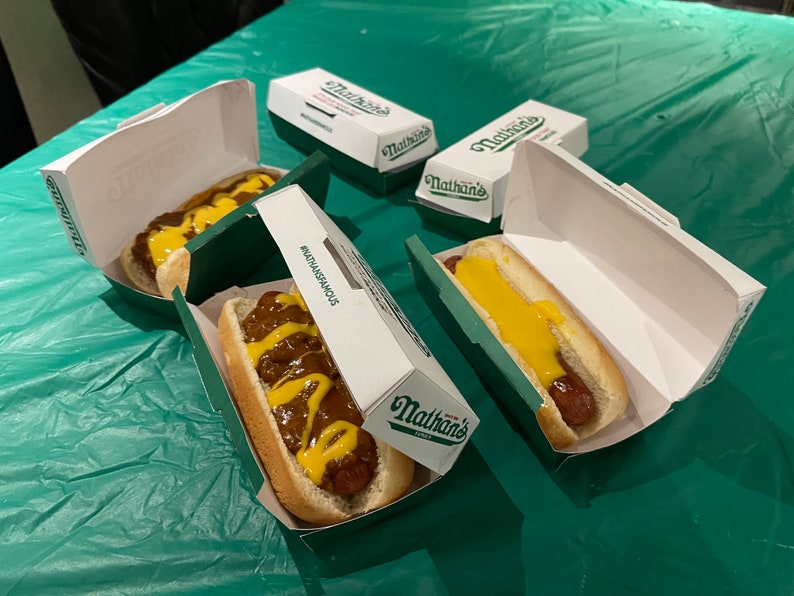 K&ouml;nnte beinhalten: Drei Nathan's Famous Hotdogs in wei&szlig;en Papierbeh&auml;ltern mit gr&uuml;nen Deckeln. Zwei Hotdogs sind mit Chili und gelbem Senf belegt. Ein Hotdog ist mit gelbem Senf belegt. Die Hotdogs befinden sich auf einer gr&uuml;nen Tischdecke.