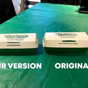 K&ouml;nnte beinhalten: Zwei wei&szlig;e und gr&uuml;ne Nathan's Famous Hotdog-Boxen nebeneinander auf einem gr&uuml;nen Tischtuch. Die Box auf der linken Seite hat den Text "IT'S OUR FOOD THAT MAKES US FAMOUS!" und den Hashtag "#NATHANSFAMOUS". Die Box auf der rechten Seite hat den Text "IT'S OUR FOOD THAT MAKES US FAMOUS" und den Hashtag "#NATHANSFAMOUS". Der Text "OUR VERSION" befindet sich unter der linken Box und der Text "ORIGINAL" unter der rechten Box.