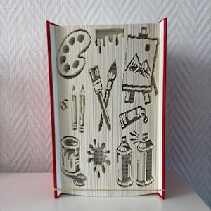 Peut inclure: Sculpture de livre avec une tranche rouge et des pages sculptées d'images liées à l'art. Les images incluent une palette de peinture, des pinceaux, des crayons, un pot de peinture, des éclaboussures de peinture, une toile et des bombes de peinture.