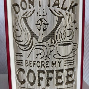 Peut inclure: Une sculpture en livre plié avec des couvertures rouges présentant le texte DON'T TALK BEFORE MY COFFEE. Le design comprend une silhouette avec un doigt sur les lèvres dans un geste de silence, à côté d'une tasse de café fumante avec des étoiles.