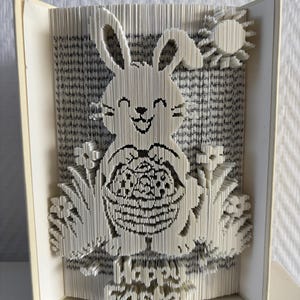 May include: A handmade book sculpture featuring a smiling bunny holding a basket, crafted from the pages of a book. The words "Happy Easter" are visible at the bottom. The bunny is surrounded by grass and a sun. The book is cream-colored.
