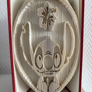 Peut inclure: Sculpture de livre fait main représentant Stitch, le personnage de Lilo & Stitch. L'œuvre est réalisée en pliant méticuleusement les pages d'un livre pour former l'image du personnage. Le livre a une couverture rouge.