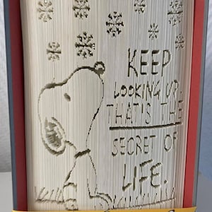 Può includere: Una scultura in libro piegato che raffigura il profilo di Snoopy che guarda verso l'alto verso fiocchi di neve che cadono. Il testo recita: KEEP LOOKING UP. THAT'S THE SECRET OF LIFE. Un nastro giallo con un motivo Snoopy avvolge la base.
