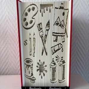 Può includere: Una scultura di libro con pagine piegate che formano figure artistiche, tra cui una tavolozza, due pennelli incrociati, un cavalletto con un paesaggio montano, matite, un tubetto di colore, un barattolo, una macchia e due bombolette spray.