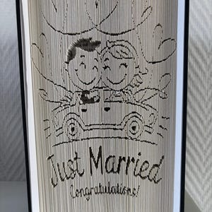 May include: A book sculpture with a wedding theme, carved to show a couple in a car, hearts, and the words "Just Married Congratulations!" The book has black covers and white pages.