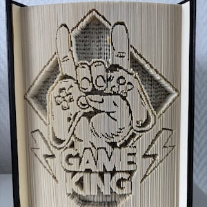 Könnte beinhalten: Ein Buch mit kunstvoll gefalteten Seiten, die das Design "Game King" zeigen. Das Kunstwerk zeigt einen Gamecontroller, Blitze und eine Raute. Das Buch hat einen schwarzen Einband und der Text ist auf den Seiten sichtbar.