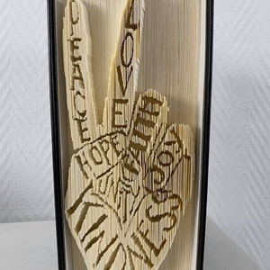 Può includere: Una scultura di arte libraria di un segno di pace realizzata piegando le pagine di un libro. Il segno di pace è composto dalle parole "Peace", "Love", "Hope", "Kindness" e "Joy".
