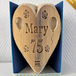 May include: A heart-shaped book art piece with the name "Mary" and the number "75" carved into the pages. The book is displayed in a blue frame, with rose and balloon designs. A ribbon with hearts decorates the base.