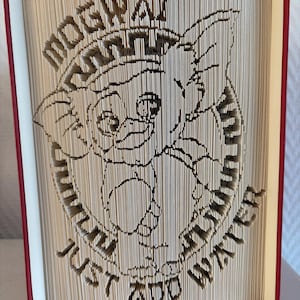 May include: A book sculpture with a red cover and white pages, featuring a cut-out design of a cartoon character with the text "Mogwai, Just Add Water". The artwork is intricately carved into the pages, creating a unique 3D effect.