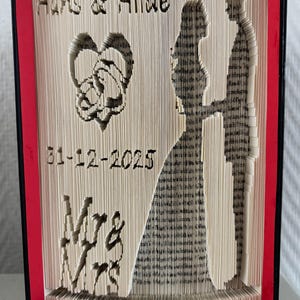 Peut inclure: Une œuvre d'art unique en forme de livre sur le thème du mariage. Les pages du livre sont pliées et coupées pour révéler un cœur, une silhouette de mariés et la date 31-12-2025. Les noms "Hans & Hilde" et "Mrs & Mrs" sont également visibles. Le livre a une bordure rouge et noire.