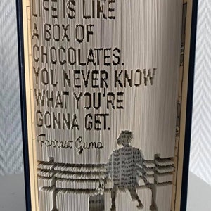 May include: A folded book art sculpture featuring the quote "LIFE IS LIKE A BOX OF CHOCOLATES. YOU NEVER KNOW WHAT YOU'RE GONNA GET." and the signature "Forrest Gump" with a silhouette of a person sitting on a bench, created by folding pages.