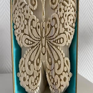 May include: A book sculpture in the shape of a butterfly, with detailed patterns cut into the pages. The butterfly is displayed in a teal box with gold edging, and a ribbon with butterfly motifs. The sculpture is a unique decorative item.