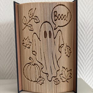 Könnte beinhalten: Eine Buchfaltkunst-Skulptur mit einem Geist, einem Kürbis und Herbstblättern. Der Text "Booo!" steht in einer Sprechblase über dem Geist. Die Seiten sind gefaltet, um diese Formen zu erzeugen, wobei dunkle Buchdeckel an den Seiten sichtbar sind.