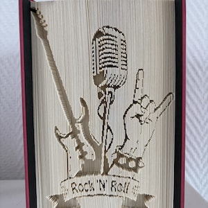 Könnte beinhalten: Ein einzigartiges Buchkunstwerk mit Rock'n'Roll-Motiv. Das Design, aus den Seiten geschnitzt, zeigt eine Gitarre, ein Mikrofon und eine Handgeste. Die Worte "Rock 'N Roll" sind auf einem Banner zu sehen. Das Buch hat einen roten Buchrücken.