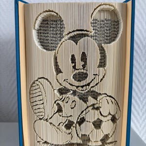 May include: A book sculpture featuring folded pages that form the shape of a cartoon mouse character holding a football. The book has blue covers and cream-coloured pages with visible text, creating a three-dimensional image of the character.