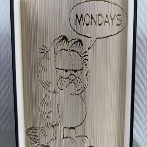 Peut inclure: Une sculpture en livre plié représentant le personnage Garfield avec une bulle contenant le texte MONDAYS. Les pages sont pliées avec précision pour créer le contour du chat et le lettrage sur les bords verticaux des pages.