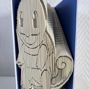 Könnte beinhalten: Eine Buchskulptur aus gefalteten Seiten, die die Form einer Zeichentrick-Eidechsenfigur mit geringeltem Schwanz bilden. Das Buch ist geöffnet und zeigt die geformten weißen Papierkanten vor einem einfarbigen blauen Hintergrund.