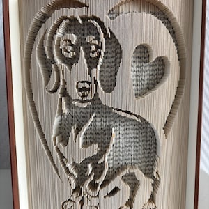 May include: A dachshund dog silhouette carved into the pages of a book, surrounded by a heart shape. The book's pages are light beige, with a brown cover. The dog's features are detailed, and the heart is open.