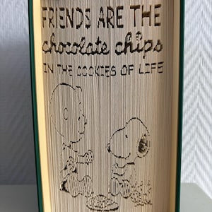 May include: A book sculpture with the text "FRIENDS ARE THE chocolate chips IN THE COOKIES OF LIFE" carved into the pages. The design features Charlie Brown and Snoopy. The book has green covers and yellow-toned pages.