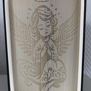 May include: A book sculpture with a carved angel design. The pages are cut and folded to create the image of an angel with wings, a halo, and a flowing gown. The book has a black cover and white pages.