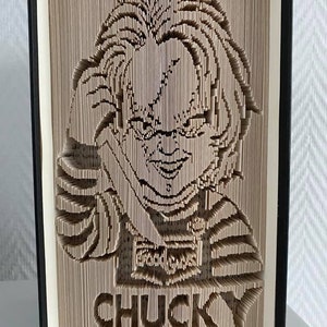 May include: A book art sculpture of the character Chucky from the Child's Play horror movie franchise. The sculpture is made from pages of a book that have been folded and cut to create a 3D image of Chucky's face and body. The sculpture is mounted on a black book cover.
