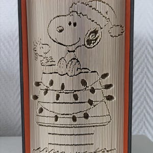 Può includere: Scultura di libro con Snoopy con cappello di Babbo Natale e Woodstock, ritagliati nelle pagine. Il design include una stringa di luci e una firma. Il libro ha il dorso nero e l'interno arancione.