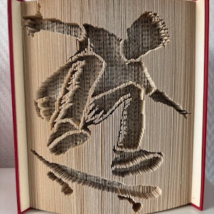 Op de afbeelding: Een gevouwen boekkunstsculptuur met het silhouet van een persoon die een skateboardtruc uitvoert. De pagina's zijn nauwkeurig gevouwen om de vorm van een skateboarder in de lucht boven een board te vormen, met rode kaften aan de zijkanten.