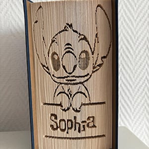 Può includere: Una scultura in libro piegato raffigurante il personaggio Stitch di Lilo e Stitch, con il nome Sophia visualizzato in un riquadro rettangolare sotto il personaggio. Le pagine del libro sono piegate con precisione per creare l'immagine e il testo.