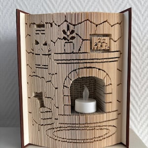 May include: A folded book art sculpture featuring a detailed fireplace scene with a mantel, a potted plant, and a small framed picture. A white battery-operated tea light candle sits inside the arched fireplace opening, creating a warm glow.