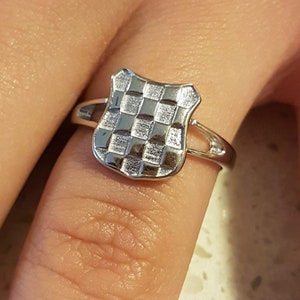 May include: A silver ring featuring a shield-shaped design with a checkered pattern. The ring is shown on a finger, with the shield element prominently displayed. The ring's band is a simple, thin design.