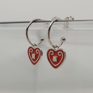 May include: A pair of silver hoop earrings with red heart charms. The hearts have a white square in the centre and a decorative design around the edge.