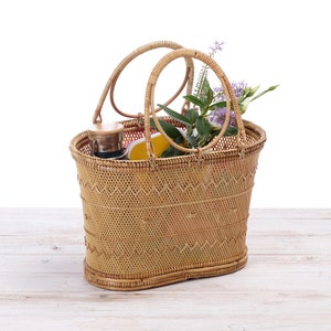 Pu&ograve; includere: Una borsa tote in rattan intrecciato con due manici arrotondati. La borsa &egrave; riempita con un thermos, un oggetto giallo e rametti di fiori viola e verdi. La borsa &egrave; appoggiata su una superficie di legno chiaro su sfondo bianco.