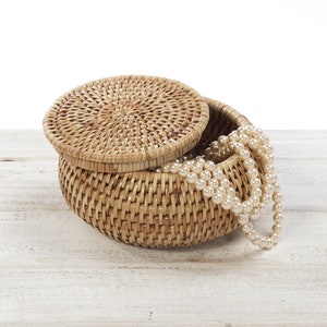 May include: A woven wicker basket with a lid, partially open, revealing a strand of white pearls inside. The basket is sitting on a white wooden surface.