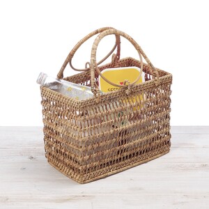 Pu&ograve; includere: Un cestino rettangolare in rattan intrecciato con due manici ad arco. All'interno del cestino ci sono una bottiglia di vetro trasparente, un contenitore giallo con la scritta "SEED" e altri oggetti. Il cestino &egrave; appoggiato su una superficie di legno bianco.