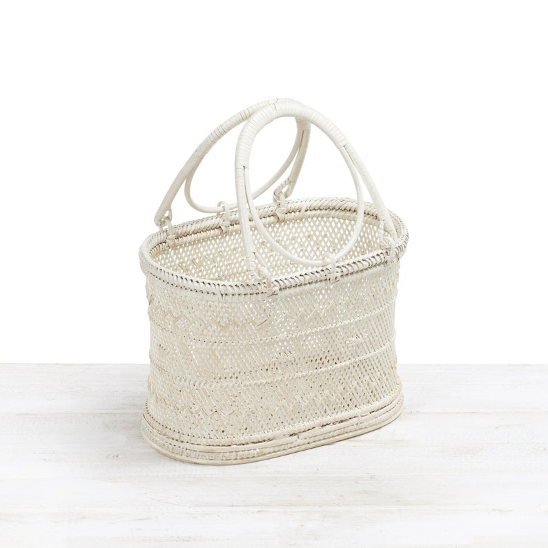 Pu&ograve; includere: Un cestino bianco intrecciato con due manici ad arco. Il cestino ha una forma ovale ed &egrave; realizzato in un materiale di colore chiaro, forse vimini o rattan. Il cestino &egrave; su una superficie di legno bianco.