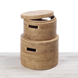 May include: Two round woven wicker baskets with lids. The top basket is open and the bottom basket is closed. The baskets are a light brown colour and have a woven pattern.