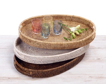May include: Three oval wicker trays stacked on top of each other. The top tray is natural wicker, the middle tray is white wicker, and the bottom tray is brown wicker. There are four small glass cups on the top tray.