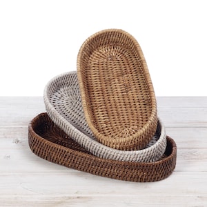 May include: Three oval woven rattan trays in shades of brown and white are stacked on a white wooden surface. The trays are empty and have a natural, textured appearance, suitable for serving or display.
